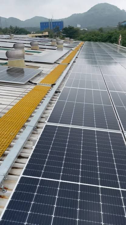 Rows of solar panels on an industrial metal roof with yellow walkways and turbine ventilators, hills in the background