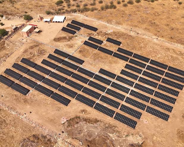 Aerial view of a fenced ground-mounted solar array laid out in rows on dry, rocky terrain